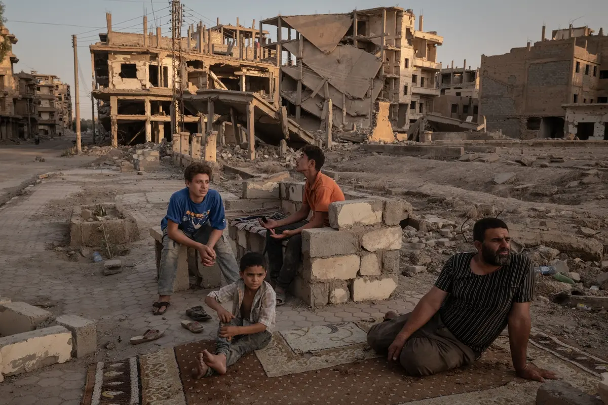 Title: A Syrian City Rebuilds, Still Divided Credit: © Nicole Tung, VII Photo, for The New York Times Caption: Abdelatif Daham Al Hummada (right) sits with his sons and nephew on the street outside their heavily damaged home, where the family often sleeps. 20 August 2025 in Deir al-Zour, Syria. Story: Long neglected by the Syrian state and one of the first cities to rise up in the 2011 revolution, Deir al-Zour endured years of siege, bombardment, and successive occupation by government forces, ISIS, and Kurdish-led fighters. The conflict left around 75% of the city’s infrastructure damaged or destroyed. In 2025, the Euphrates River marked a divide; the government controlled one bank, the Kurdish-led Syrian Democratic Forces (SDF) the other, complicating daily movement, trade, and access to services. For those who remained, and those who returned, rebuilding continued regardless.